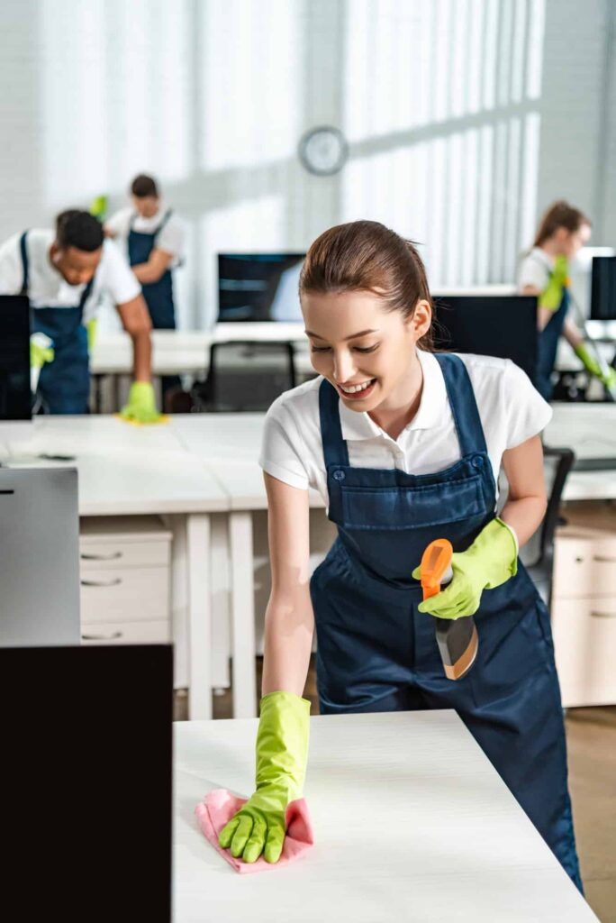 Cheerful Cleaner In Overalls Cleaning Office Desk With Rag 1.jpg